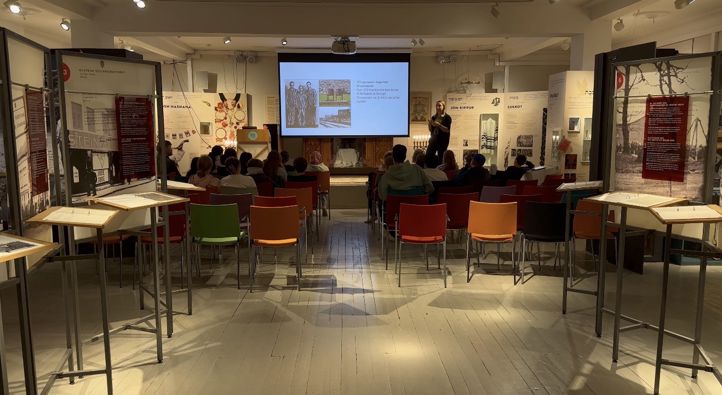 en gruppe mennesker som sitter ved bord i et rom med en stor skjerm en gruppe mennesker som sitter ved bord i et rom med en stor skjerm