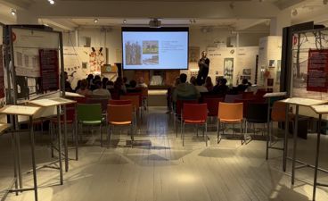 en gruppe mennesker som sitter ved bord i et rom med en stor skjerm en gruppe mennesker som sitter ved bord i et rom med en stor skjerm