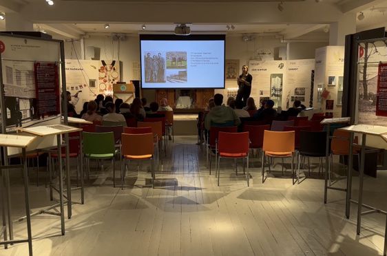 en gruppe mennesker som sitter ved bord i et rom med en stor skjerm en gruppe mennesker som sitter ved bord i et rom med en stor skjerm